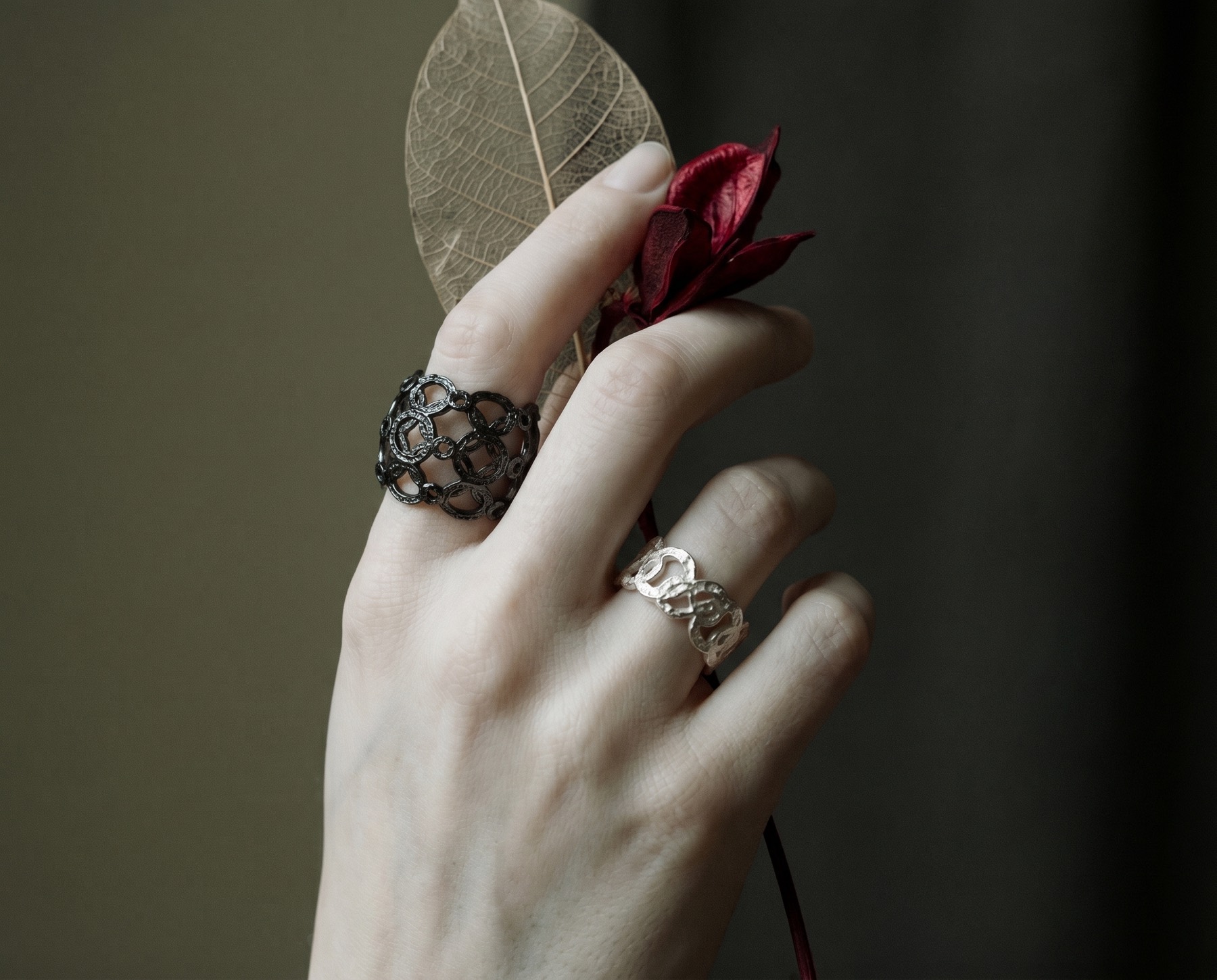 A hand wearing two handmade rings, holding a skeleton leaf and a red flower.